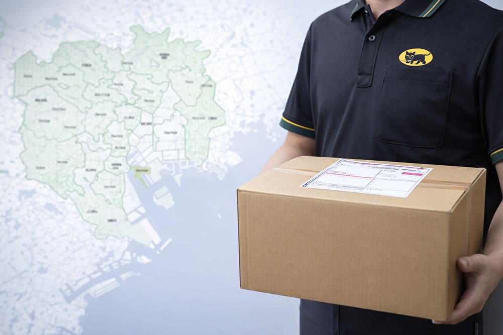 配送用の段ボールを手に持つ手元と、背景にぼんやり表示された東京都の地図イメージ。礼服・喪服が翌日到着しやすいことを連想させる。